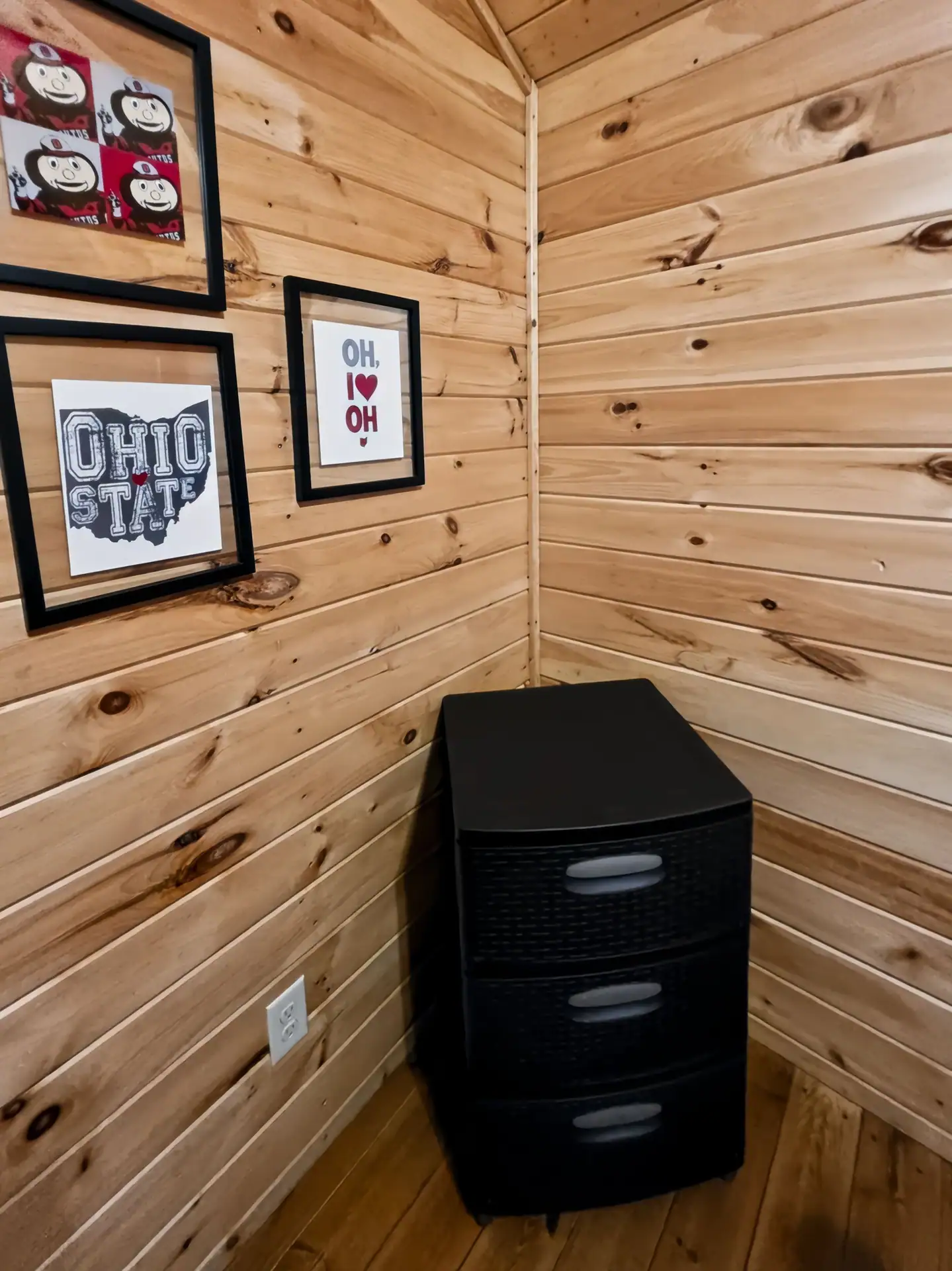 Pine-paneled bedroom corner with Ohio State framed prints and a dresser at Buckeye Hut cabin