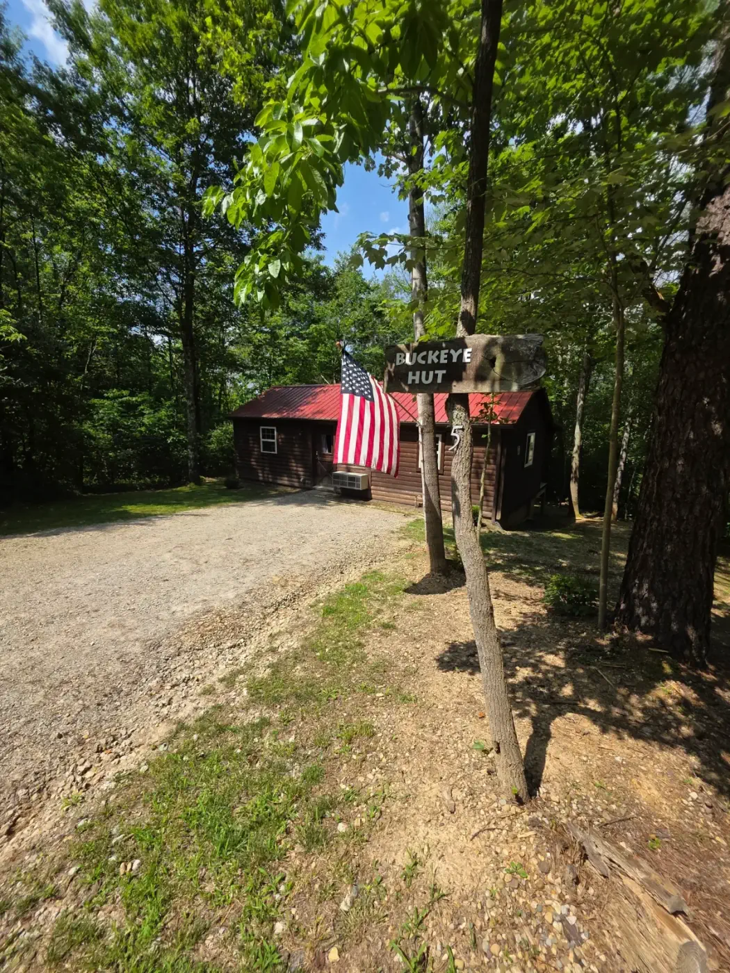View up gravel driveway to Buckeye Hut cabin with American flag and forest backdrop
