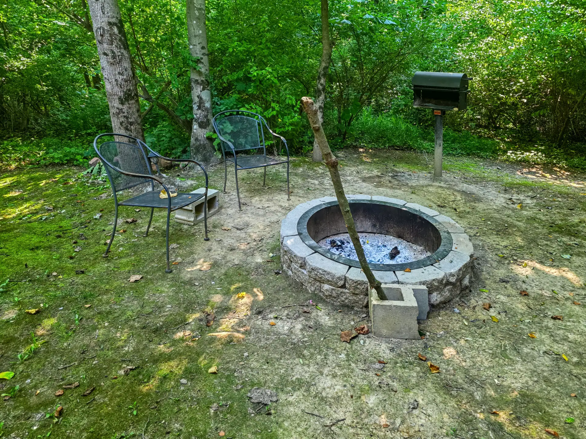Stone fire pit with metal chairs and charcoal grill in the wooded yard at Buckeye Hut cabin