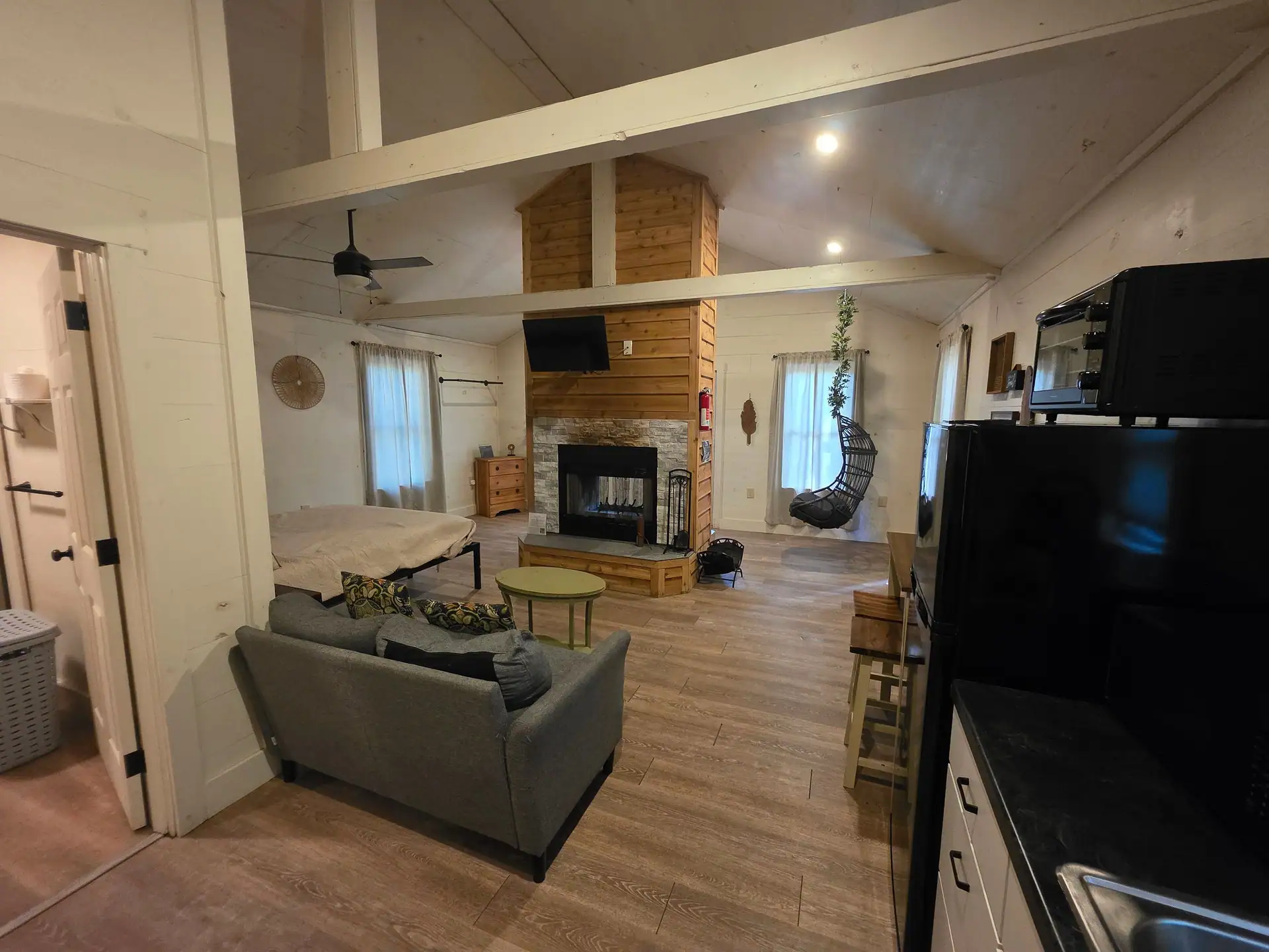 Open-plan interior with sofa, double-sided fireplace, hanging egg chair and queen bed under the vaulted ceiling at Hummingbird cabin