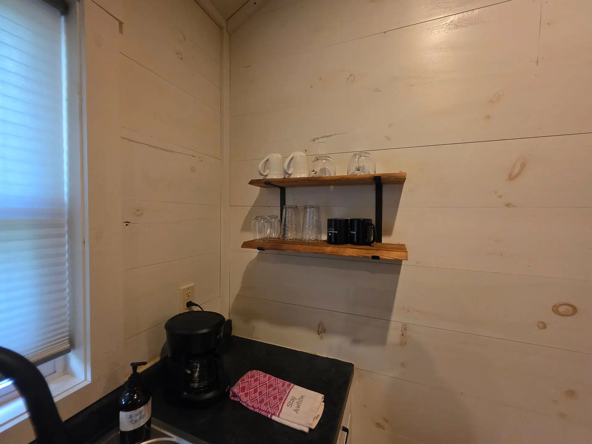 Coffee maker on the counter below open wooden shelves with mugs and glasses at Hummingbird cabin
