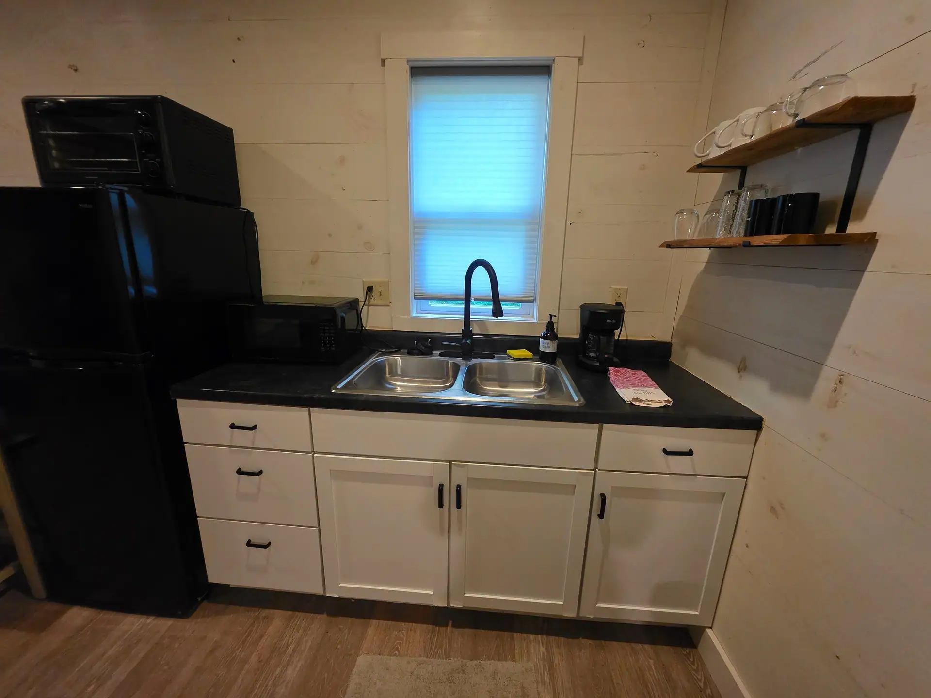 Kitchenette with white cabinets, double sink, black fridge, microwave and open shelves at Hummingbird cabin