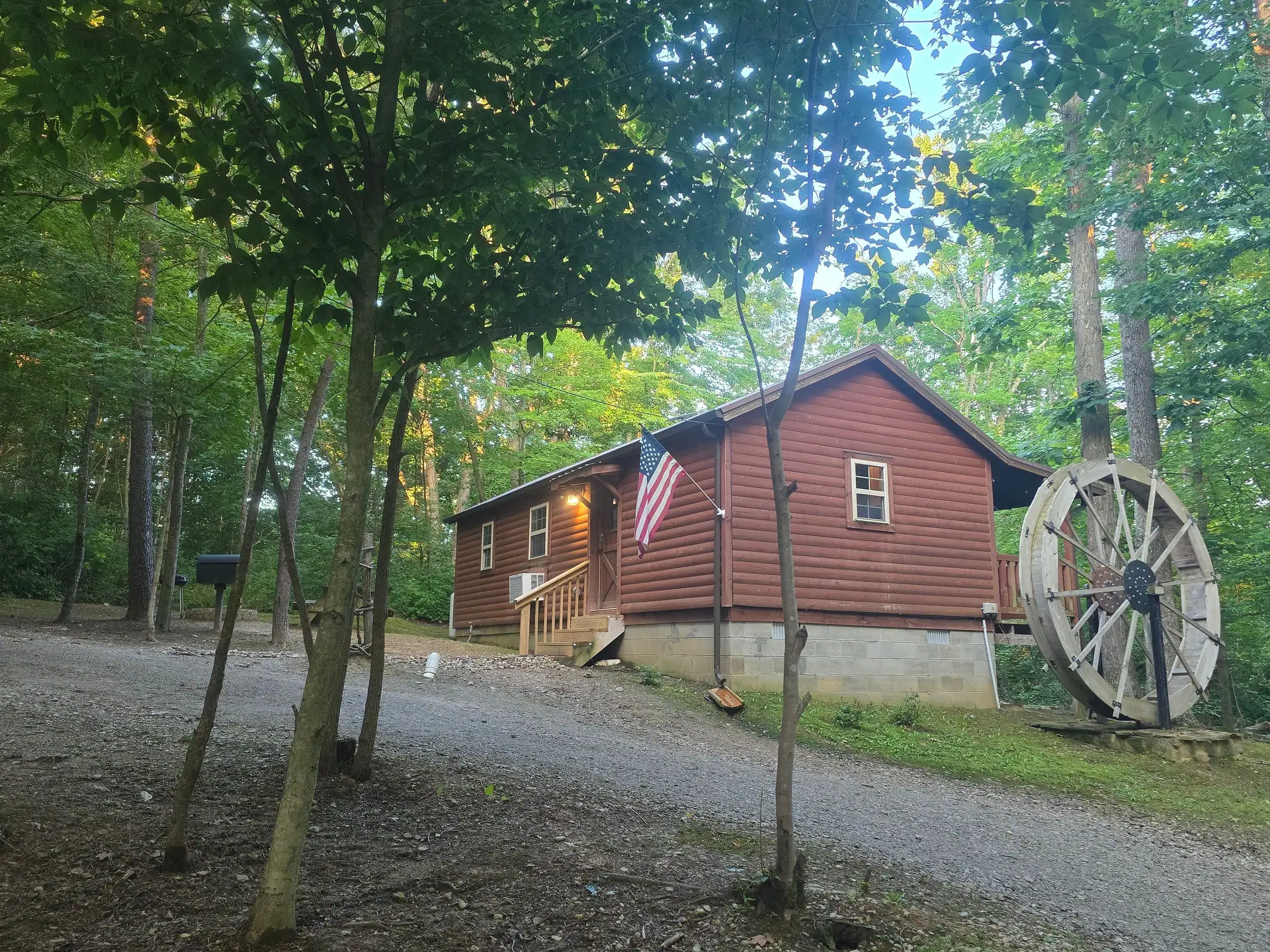 Side view of Millers Den cabin with porch light, flag and water wheel in the evening