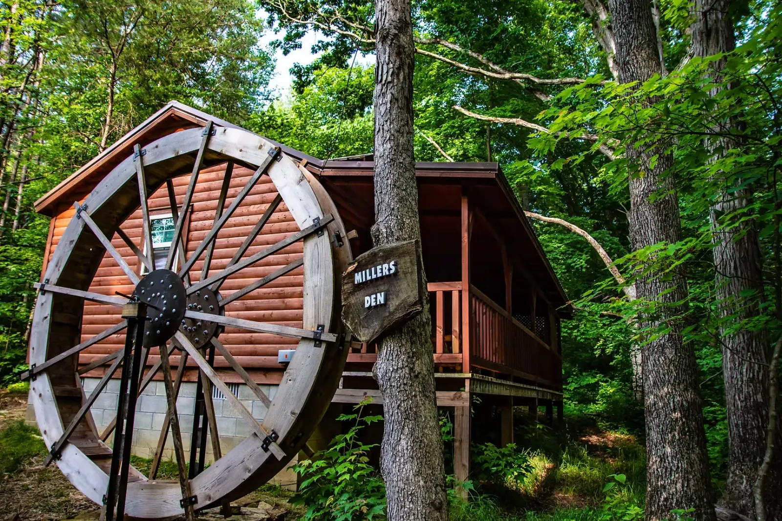 Millers Den cabin front exterior featuring large water wheel and name sign among trees