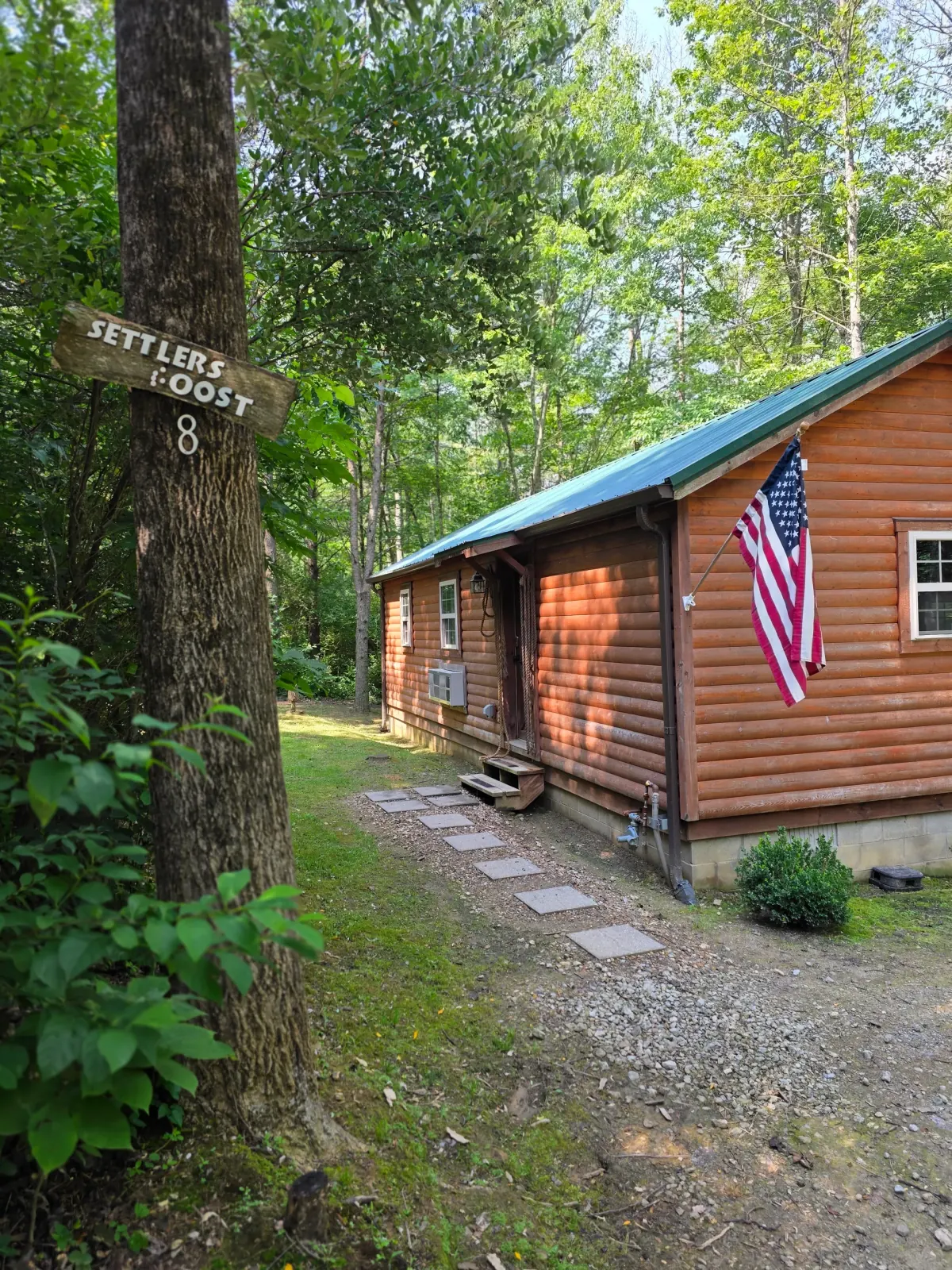 Settlers Roost cabin entrance with wooden name sign on tree, covered porch, and American flag