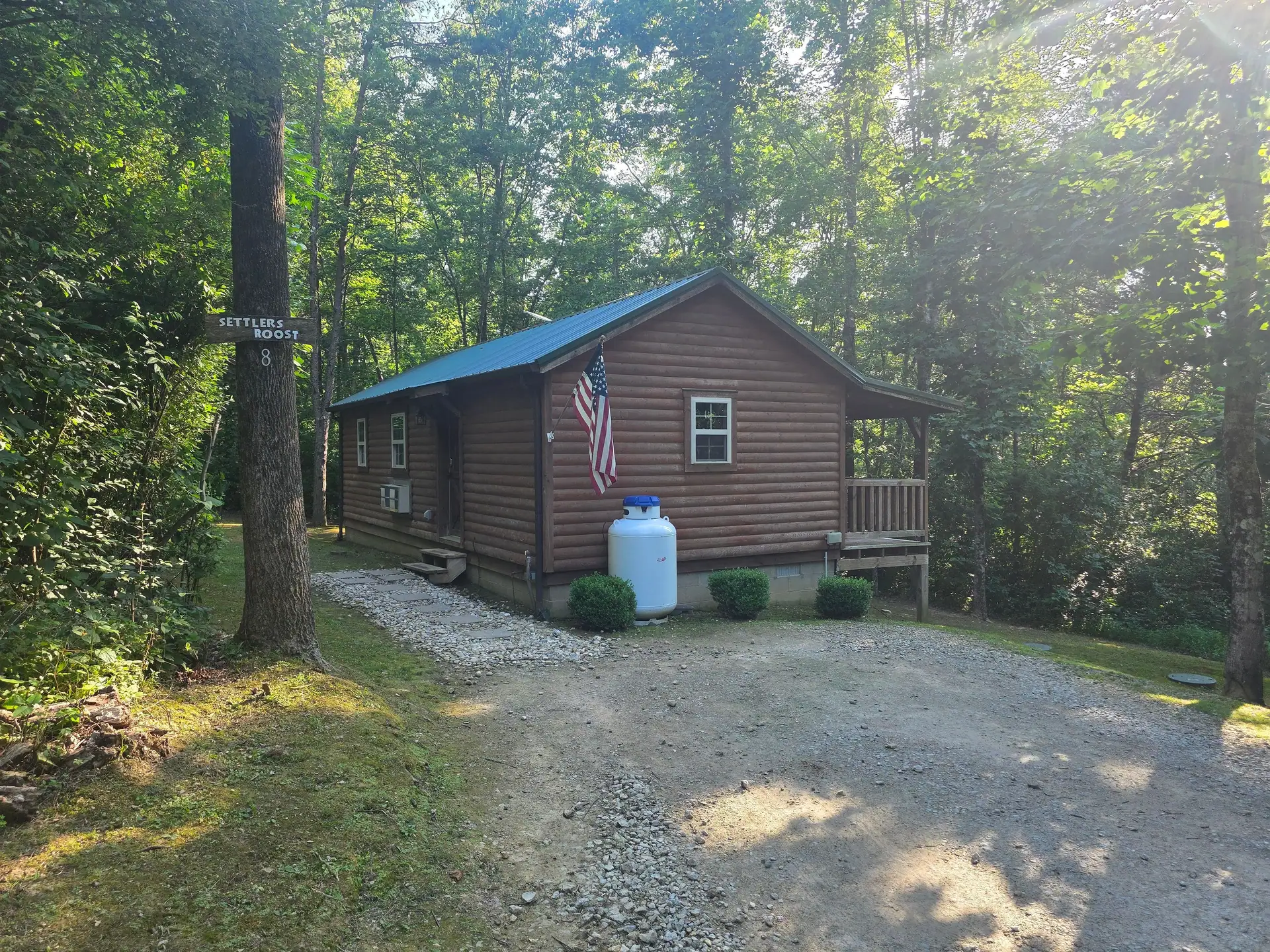Settlers Roost log cabin with green roof, front porch and gravel driveway in summer