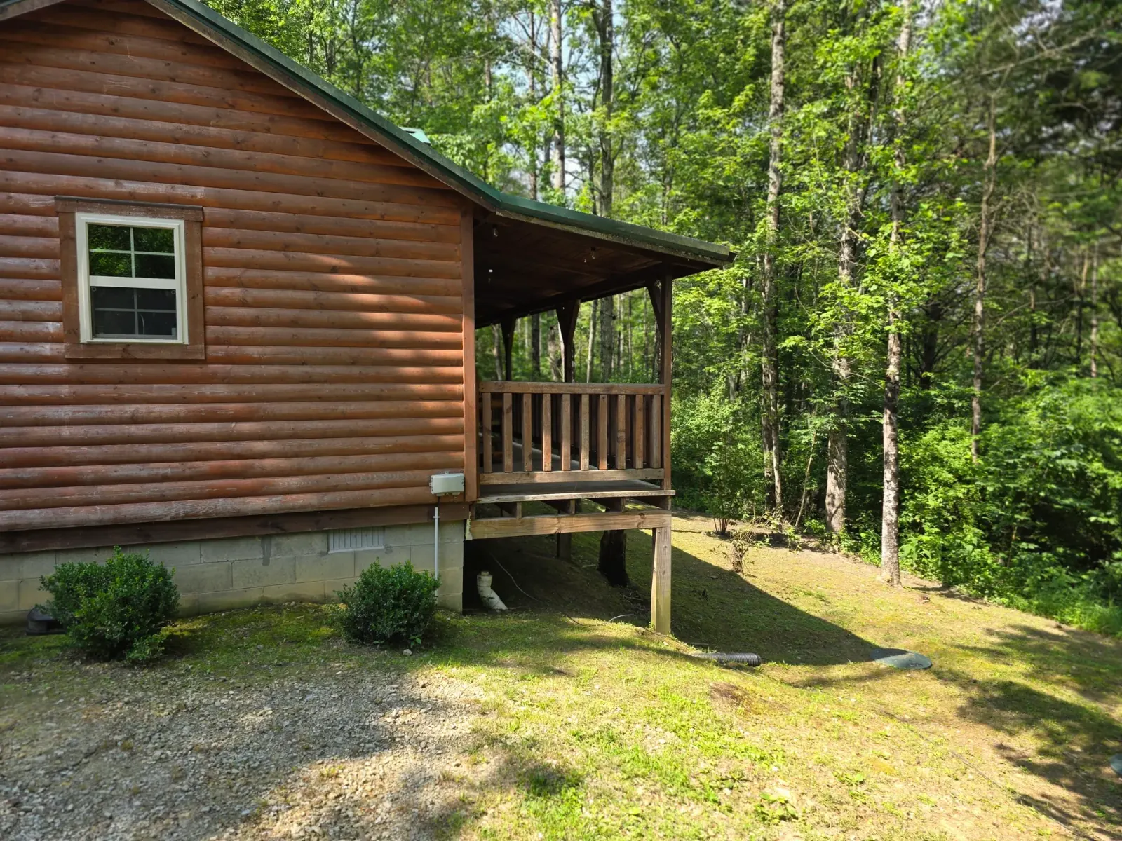 Side view of Settlers Roost log cabin showing covered porch and green metal roof surrounded by woodland